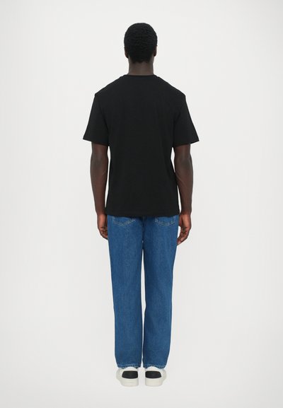 T-shirt en coton noir à manches courtes, associé à un jean en denim bleu et des baskets blanches avec des accents noirs ; vue de dos.