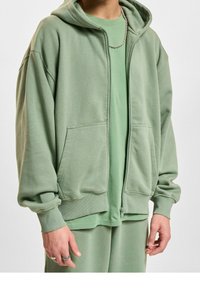 Groene zip-up hoodie met grote voormouwen, geribde manchetten en een gladde textuur, gelaagd over een bijpassend t-shirt.