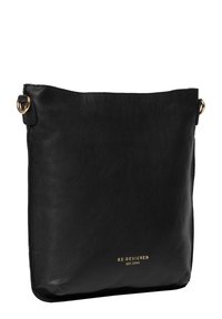 Bolso cruzado de piel negra con una textura suave, presentando un logo dorado en relieve y un diseño simple y minimalista. Forma grande y cuadrada.