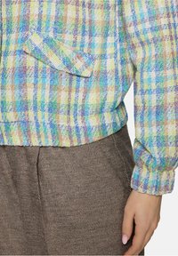 Wielokolorowa, tkana kurtka w pastelowych odcieniach niebieskiego, zielonego, żółtego i brązowego z fakturowanym wykończeniem i detalem w postaci naszywanej kieszeni; w zestawie z brązowymi spodniami.