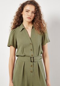 Olivengrüner Jumpsuit aus leichtem Stoff, kurze Ärmel, durchgehende Knopfleiste, tailliert mit einem Gürtel, mit braunen Knöpfen und einem Kragen.