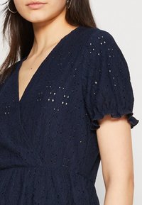 Mörkblå klänning med v-ringning, korta puffärmar med volangkanter och ögla-textur med små diamantmönster genom hela.