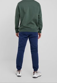 Grön sweatshirt med rund halsringning, tillsammans med marinblå sweatpants. Sweatshirten har ribbade ärmslut och fåll. Vita sneakers med grå och orange detaljer.