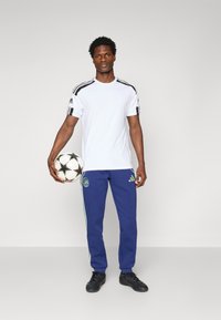 Hvit sporty t-skjorte med svarte striper på skuldrene, kombinert med blå joggebukser med grønne detaljer og logo, holder en fotball.