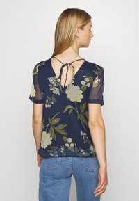 Blusa naval floral com mangas bufantes, um laço nas costas e um decote em V profundo. O tecido é leve, com grandes padrões de flores creme e verdes.
