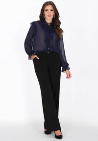 Tengerészkék áttetsző blúz fodros nyakkivágással és ujjakon, fekete magasderekú nadrággal párosítva. A modell fekete magassarkút visel, kezei a zsebekben.