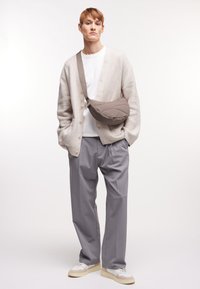 Borsa a tracolla in taupe, indossata a tracolla. Modello in cardigan crema sopra una t-shirt bianca e pantaloni plissettati grigio chiaro, con sneaker bianche.