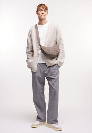 Borsa a tracolla in taupe, indossata a tracolla. Modello in cardigan crema sopra una t-shirt bianca e pantaloni plissettati grigio chiaro, con sneaker bianche.