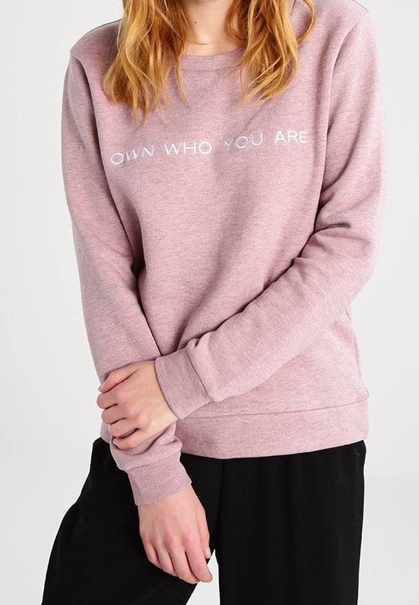 Rosa sweatshirt med en mjuk textur, har texten "ÄG DEN DU ÄR" i vitt över bröstet, i kombination med svarta byxor.