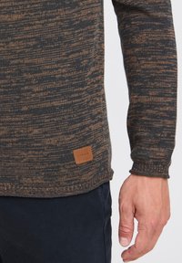 Gestrickter Pullover in Dunkelgrau mit braunen Sprenkeln. Verfügt über gerippte Bündchen und eine strukturierte Oberfläche. Etikette mit Branding an der Seite.