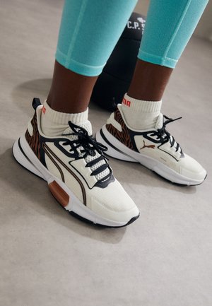 Persona con zapatillas blancas y negras con rayas de tigre y calcetines blancos Puma de pie sobre un suelo gris cerca de un peso negro.