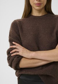 Brūns adīts džemperis ar teksturētu apdari, brīvs piegriezums un ruļļveida piedurknes. Mīksts materiāls brīvi krīt ap ķermeni.