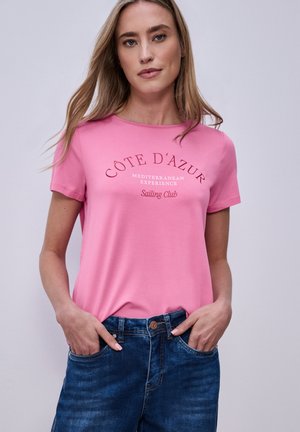 Frau trägt ein pinkes T-Shirt mit dem Schriftzug "Côte D'Azur Mediterranean Experience Sailing Club", kombiniert mit blauen Jeans, Hände in den Taschen, neutraler Hintergrund.