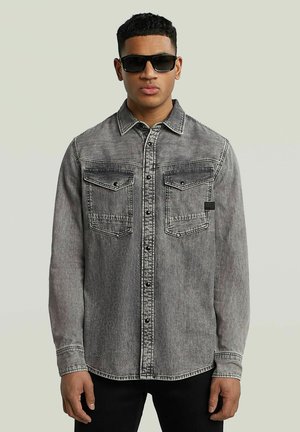 Grå denimskjorta med två framfickor, knapplåsdesign, kontrasterande sömmar och en rundad nederkant. Modellen bär svarta solglasögon.