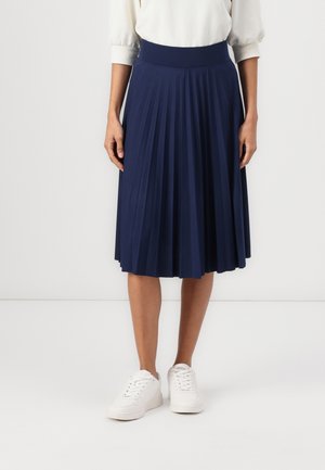 Personne portant une jupe midi plissée bleu marine, un chemisier blanc à manches longues et des baskets blanches, debout devant un fond uni.