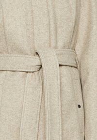 Cappotto di lana beige chiaro con cintura, cuciture in vista e superficie testurizzata. Include un dettaglio a scatto sul lato.