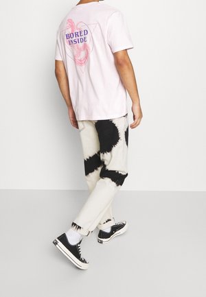 Roze katoenen T-shirt met de tekst "BORED INSIDE" op de rug, gecombineerd met crèmekleurige jeans met zwart tie-dye, en zwarte sneakers met witte accenten.