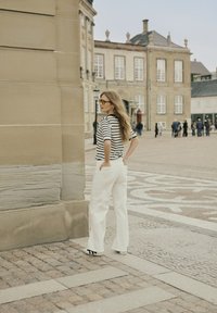 Striped black and white short-sleeve top, white high-waisted wide-leg pants, sunglasses, with long wavy hair, standing near a stone wall.