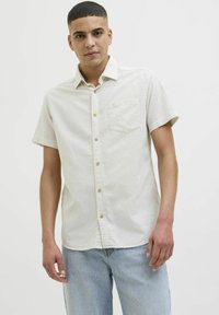 Chemise à manches courtes et col, en tissu léger couleur crème avec un motif de petits points subtils. Comprend une poche avant et une fermeture à boutons.
