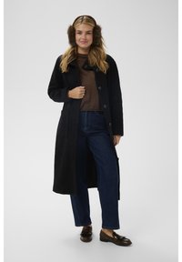 Donna che indossa paraorecchie marroni, cappotto lungo nero, maglione marrone, jeans blu e mocassini marroni, in piedi davanti a uno sfondo semplice.