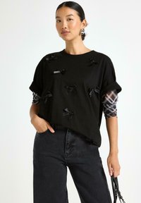 Kvinde iført en sort oversized t-shirt med små sorte sløjfer, ternede langærmede skjorter under, sorte jeans, sølv hoop øreringe og holder en taske.