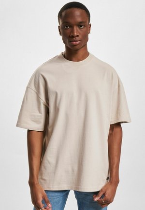 T-shirt beige oversize in cotone con maniche corte, scollatura rotonda e un piccolo logo accentato vicino all'orlo. Abbinato a jeans azzurri chiari.