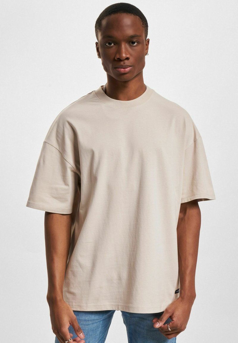 T-shirt beige oversize in cotone con maniche corte, scollatura rotonda e un piccolo logo accentato vicino all'orlo. Abbinato a jeans azzurri chiari.