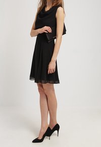 Vestido negro sin mangas con escote drapeado, falda acampanada y dobladillo transparente. La modelo sostiene un bolso negro de mano y lleva puestos unos tacones altos negros.