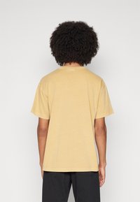 Camiseta de algodón en color mostaza con cuello redondo y mangas cortas, que presenta un corte relajado y un simple detalle de costura.