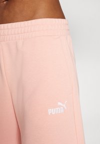 Gaiši rozā treniņbikses, izgatavotas no mīksta auduma, ar elastīgu jostu un baltu izšuvu Puma logo kreisajā pusē.