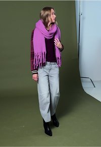 Ternet jakke med et lilla frynset tørklæde, mørk skjorte, lysegrå vide jeans, og sorte spidse ankelstøvler på en grøn baggrund.