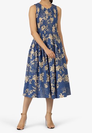 Mouwloze blauwe midi-jurk met beige bloemenprint, gerimpeld lijfje, geplooide rok, gedragen met sandaal met doorzichtige hak en enkelbandje.