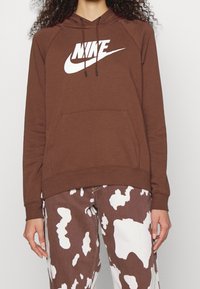Brown hoodie with a white Nike logo, featuring a kangaroo pocket, drawstring hood, and ribbed cuffs. Paired with patterned brown and white jeans.