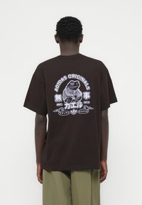 Καφέ βαμβακερό μπλουζάκι με εκτυπωμένη εικόνα ενός βατράχου με φούτερ, με το κείμενο "ADIDAS ORIGINALS" πάνω και επιπλέον ιαπωνικούς χαρακτήρες.