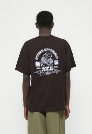 Brūna kokvilnas T-krekls ar grafisku attēlu - vardzēnis ar kapuci, augšpusē teksts "ADIDAS ORIGINALS" un papildu japāņu rakstzīmes.