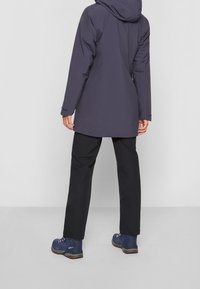 Vattentålig mörk lila jacka med huva, kombinerad med svarta byxor och blå vandringskängor. Har en enkel och funktionell design.