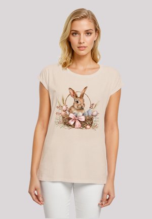 Beige Baumwoll-T-Shirt mit einem Hasen in einem Nest mit Ostereiern und einer pinken Schleife, runder Halsausschnitt und kurzen Ärmeln. Glatte Textur.