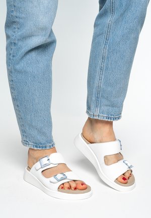 Persona con vaqueros azul claro y sandalias blancas con dos hebillas ajustables, de pie sobre un fondo blanco.