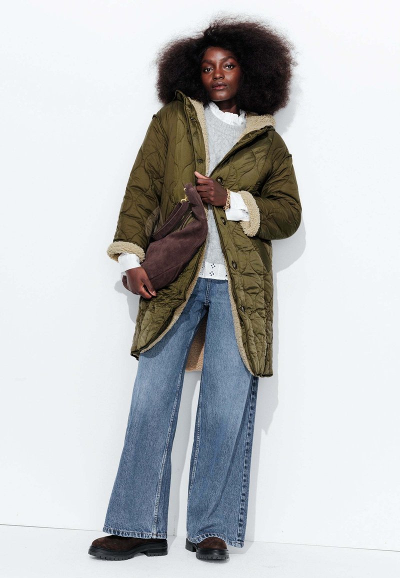 Manteau matelassé vert olive avec doublure beige, associé à un pull gris et un jean bleu à larges jambes. Le mannequin tient un sac en daim marron.