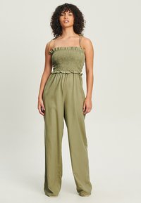 Combinaison couleur olive avec un corsage smocké, des volants, des bretelles ajustables fines et des jambes larges en tissu léger.