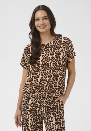 Usmievajúca sa žena s dlhými tmavými vlasmi v rovnakom voľnom leopardom vzorovanom tričku a nohaviciach, s jednou rukou v vrecku, na jednofarebnom pozadí.