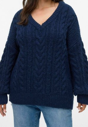 Femme portant un pull en grosse maille bleu foncé à col en V avec des motifs torsadés et un jean en denim bleu clair sur fond uni.