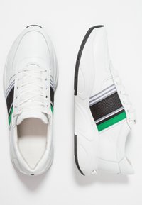 Białe sneakersy z siateczkowymi panelami, czarnymi i zielonymi paskami, teksturowanymi akcentami oraz amortyzowaną podeszwą. Widoczne sznurówki i logo na języku.