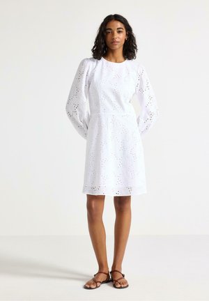 Femme aux cheveux bouclés portant une robe blanche à manches longues en broderie anglaise et des sandales plates marron, debout devant un fond clair uni.