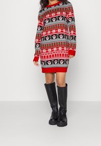 Robe-pull rouge en maille avec des motifs festifs en noir, blanc, vert et rouge. Portée avec des bottes noires montantes jusqu'au genou.