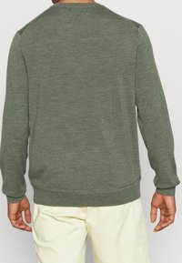 Man i olivgrön långärmad tröja och ljusbeiga byxor, vänd bort från kameran mot en ljus, enfärgad bakgrund.