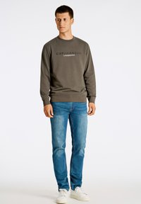 Sudadera verde oscuro con cuello redondo y logo en la parte delantera, combinada con jeans claros y zapatillas blancas, de pie frente a un fondo liso.