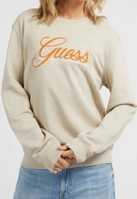Sweat-shirt beige en coton, arborant un logo "Guess" orange brodé avec "EST. 1981" en dessous. Manches avec poignets et ourlet côtelé.