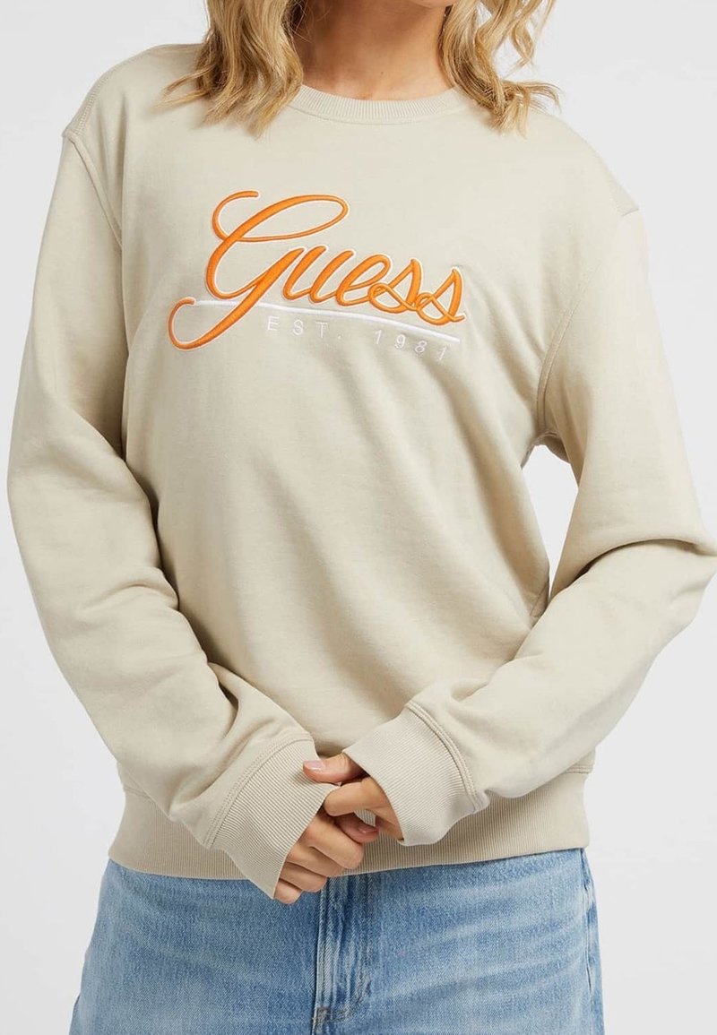 Sweat-shirt beige en coton, arborant un logo "Guess" orange brodé avec "EST. 1981" en dessous. Manches avec poignets et ourlet côtelé.