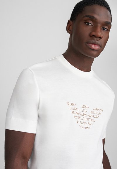 Λευκό κοντομάνικο μπλουζάκι με υφασμάτινο σχέδιο λογότυπου σε αποχρώσεις μπεζ και καφέ στο στήθος. Φτιαγμένο από ένα απαλό, ελαφρύ ύφασμα.